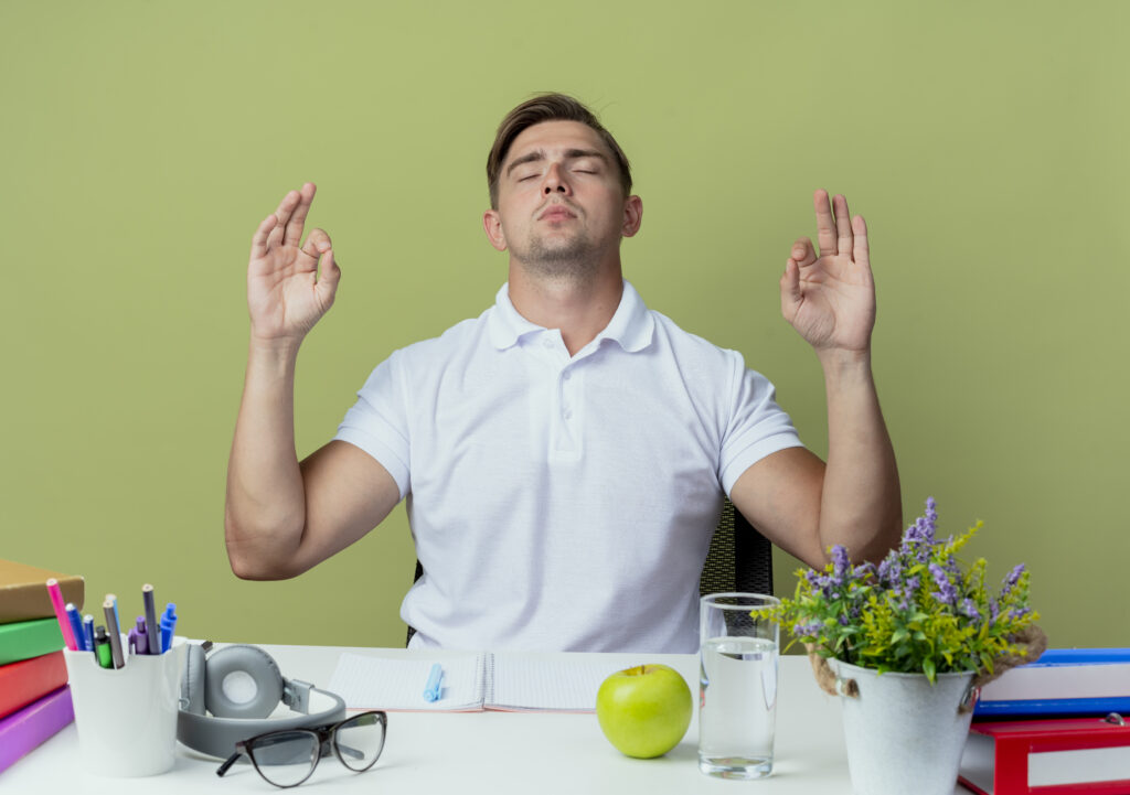 with-closed-eyes-young-handsome-male-student-sitting-desk-with-school-tools-showing-meditation-gesture-isolated-olive-green